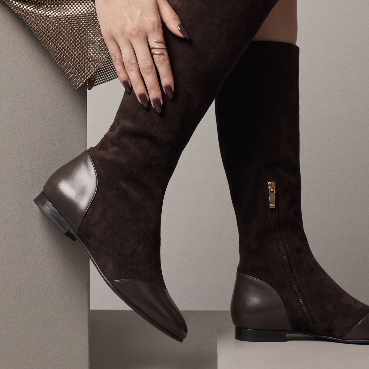 A close-up of a person wearing dark brown suede over-the-knee boots with smooth leather heel and toe panels, posed against a neutral gray background.