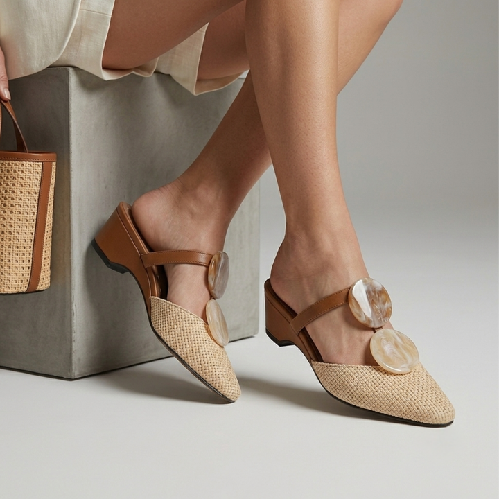 Paloma Mule in Sable Doré. A close-up of a person wearing woven tan slingback mule flats with large round shell-like embellishments on the straps, seated on a concrete block with a matching woven handbag partially visible.