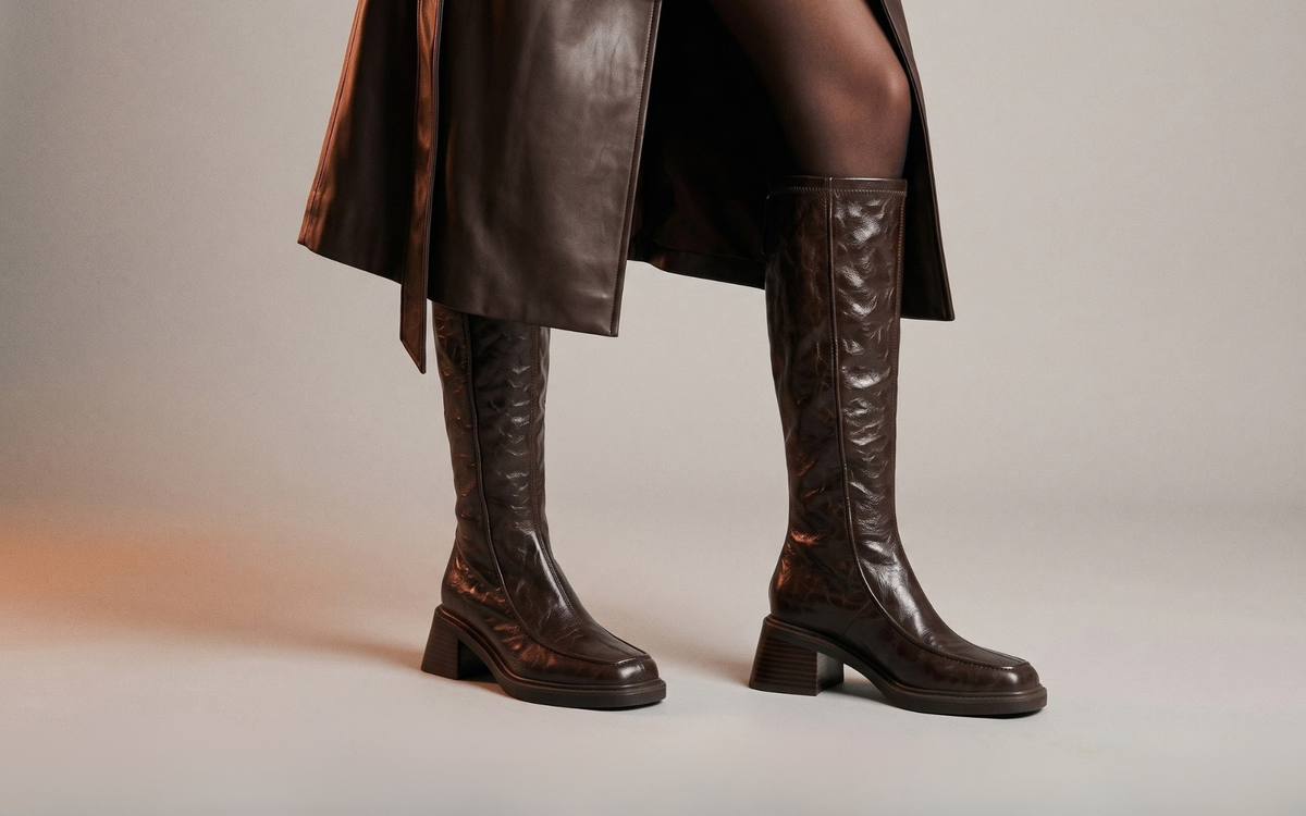 A person wearing dark brown knee-high leather boots with a low block heel and textured embossed pattern, shown from the mid-thigh down against a neutral studio background.