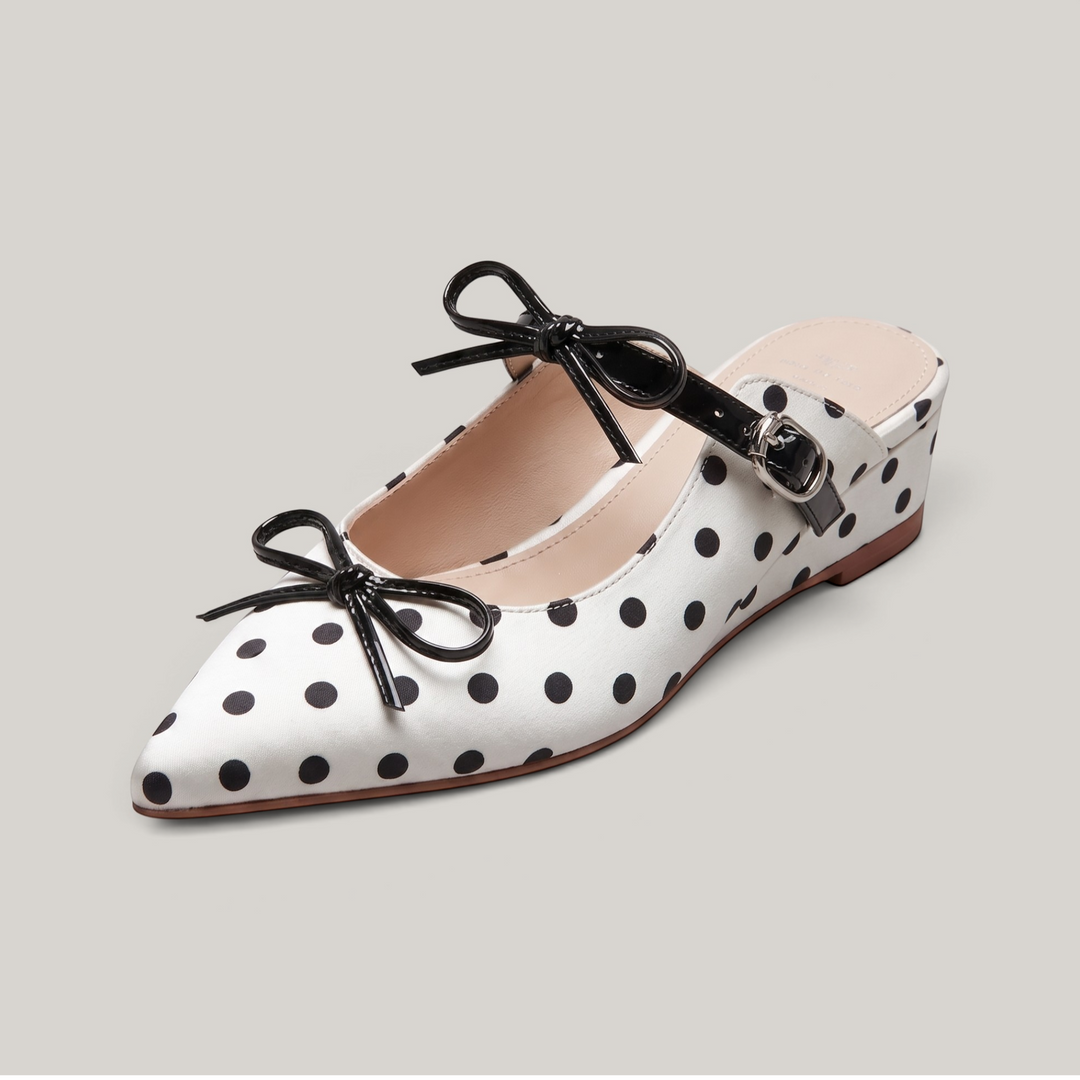 USS® Shoes Elise pointed-toe mule in white leather with black polka dots, featuring black bow accents and buckle strap.